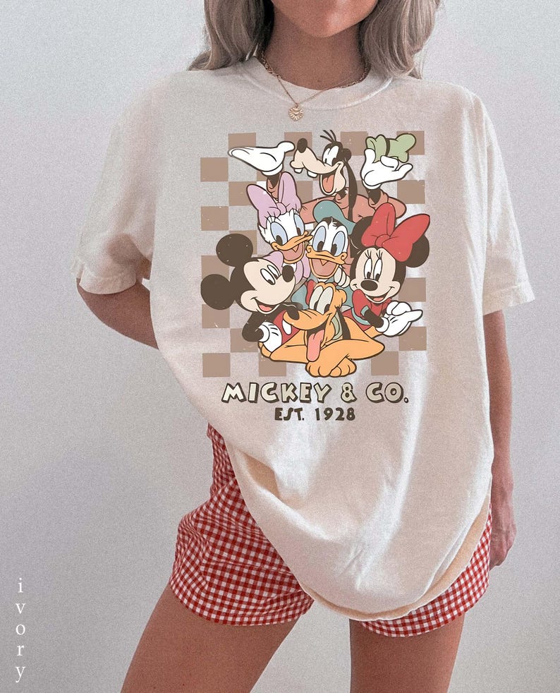 May include: Ivory t-shirt with a graphic of Mickey Mouse, Minnie Mouse, Donald Duck, Daisy Duck, Goofy, and Pluto. The design is set against a brown checkered background, with "Mickey & Co. Est. 1928" printed below. The shirt is paired with red and white checkered shorts.