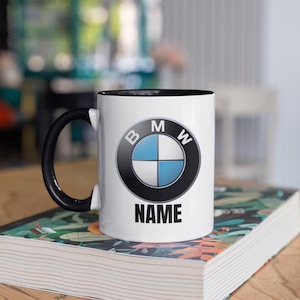 Op de afbeelding: Witte keramische mok met een zwarte binnenkant en handvat. De mok toont het BMW-logo en het woord "NAME" eronder. De mok staat op een boek met een bloemenpatroon.