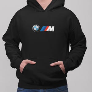 Puede incluir: Una sudadera negra con capucha que presenta el logotipo de BMW M en el centro del pecho. El logotipo consiste en el emblema circular azul y blanco de BMW seguido de la letra M en blanco, junto a tres franjas inclinadas en azul, violeta y rojo.