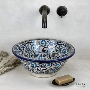 Puede incluir: Un lavabo decorativo de cerámica con un diseño floral azul y blanco. El lavabo tiene un borde azul oscuro y está colocado contra una pared blanca con un grifo y una válvula negros. Una pastilla de jabón descansa sobre un plato pequeño.