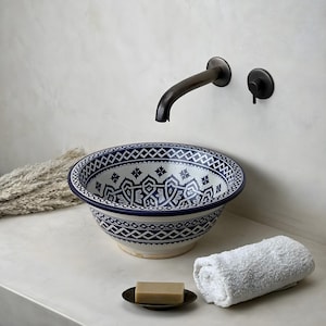 Puede incluir: Un lavabo de cerámica decorativo con un patrón geométrico azul y blanco. Un grifo de bronce oscuro está montado en la pared sobre el lavabo. Una toalla blanca enrollada, jabón y un jabonera están en la encimera. El lavabo está etiquetado "COHEN Moroccan".