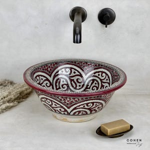 Puede incluir: Un lavabo decorativo de cerámica con un diseño estampado en rojo y blanco. El lavabo es redondo con un borde ancho y se apoya sobre una superficie blanca. Una pastilla de jabón descansa sobre un pequeño jabonera de metal oscuro. Un grifo y manija de metal oscuro están montados en la pared.