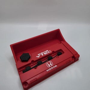 May include: A red 3D-printed Honda engine cover with the text "DOHC VTEC" and the Honda logo. The cover has a rectangular shape with a black cap and black detailing. The background is plain white.