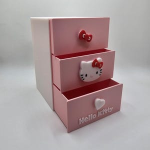 May include: A three-drawer storage unit with a white base and pink drawers. Each drawer features a decorative element: a bow, a Hello Kitty face, and a heart. The words "Hello Kitty" are embossed on the bottom drawer.
