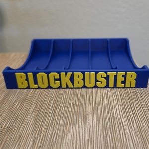 Peut inclure: Un support de cartouches de jeux vid&eacute;o Blockbuster bleu et jaune. Le support a un design incurv&eacute; avec plusieurs fentes pour les cartouches de jeu. Le mot "BLOCKBUSTER" est imprim&eacute; en lettres jaunes.