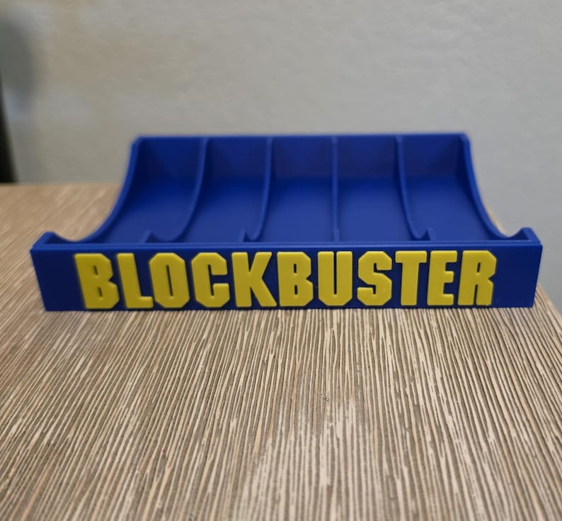 Peut inclure: Un support de cartouches de jeux vid&eacute;o Blockbuster bleu et jaune. Le support a un design incurv&eacute; avec plusieurs fentes pour les cartouches de jeu. Le mot "BLOCKBUSTER" est imprim&eacute; en lettres jaunes.