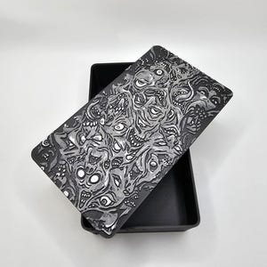 May include: A black box with a lid featuring a white and gray graphic print of a swirling pattern of eyes and faces. The box is made of a smooth, matte material.