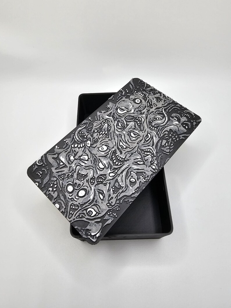 May include: A black box with a lid featuring a white and gray graphic print of a swirling pattern of eyes and faces. The box is made of a smooth, matte material.