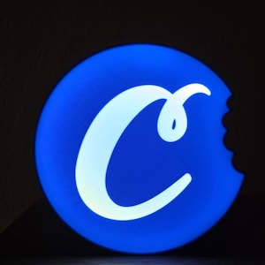 May include: A round, illuminated sign with a bright blue background and a white "C" logo. The logo has a bite taken out of the right side, suggesting a cookie. The sign is likely for a business.