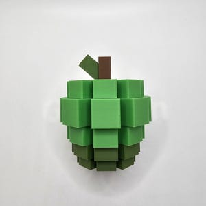 May include: A 3D-printed apple sculpture composed of stacked green and brown square blocks. The apple's body is a gradient of green shades, with a brown stem and a green leaf. The design is geometric and modern.