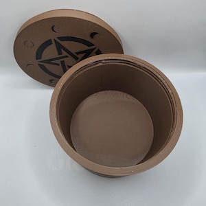 May include: A brown, round container with a lid. The lid features a black pentagram design with crescent moon accents. The container is open, revealing its interior. The item is likely used for storage or display.