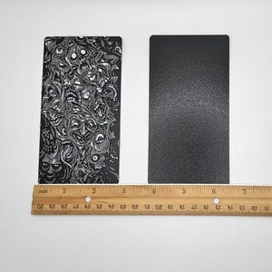 May include: Two rectangular pieces of black material, one with a white and gray pattern of faces and the other solid black. A ruler is placed below the two pieces of material.