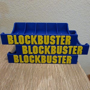 Peut inclure: Trois supports bleus et jaunes pour cartouches de jeux vid&eacute;o Blockbuster empil&eacute;s. Le mot "BLOCKBUSTER" est imprim&eacute; en jaune sur chaque support. Les supports sont sur une surface en bois brun clair.
