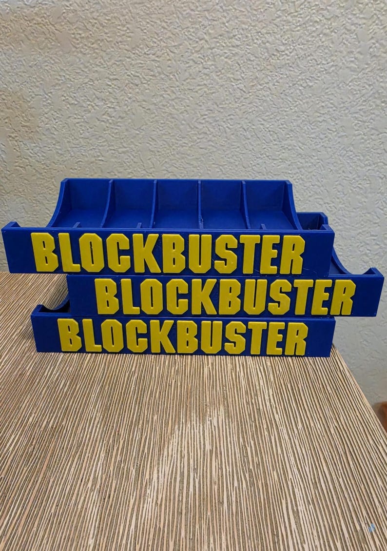 Peut inclure: Trois supports bleus et jaunes pour cartouches de jeux vid&eacute;o Blockbuster empil&eacute;s. Le mot "BLOCKBUSTER" est imprim&eacute; en jaune sur chaque support. Les supports sont sur une surface en bois brun clair.