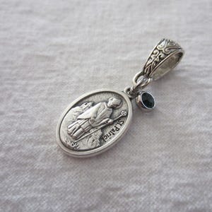 May include: An oval silver pendant featuring a relief image of St. Patrick holding a staff, with the text St. Patrick inscribed on the surface. The pendant includes a small, round, dark black gemstone charm and a decorative silver bail for attachment.