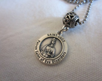May include: A silver pendant necklace featuring a circular charm with the image of Saint Lucy, the patron saint of eye disorders. The charm is inscribed with "Saint Lucy" and "Patron Saint of Eye Disorders". The necklace has a ball chain.
