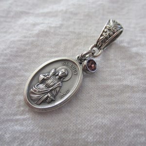 May include: An oval silver-toned pendant featuring a relief image of St. Lucy with the text ST. LUCY and ITALY. The pendant includes a small, round, dark red gemstone charm attached to a decorative silver-toned bail with an intricate leaf pattern.