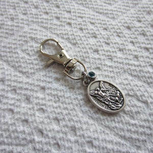 May include: A silver-toned metal keychain featuring a lobster clasp and an oval-shaped pendant depicting St. Michael. The pendant includes the text St. Michael and Italy, accented by a small, round, blue crystal bead attached to the jump ring.