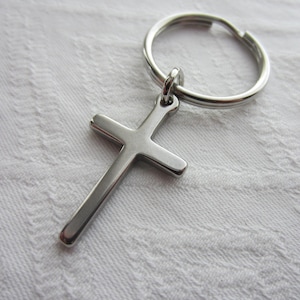 May include: A silver-toned metal cross pendant attached to a circular split key ring. The cross features a smooth, polished surface with rounded edges and a simple, clean design. The key ring is a standard round metal loop for holding keys.