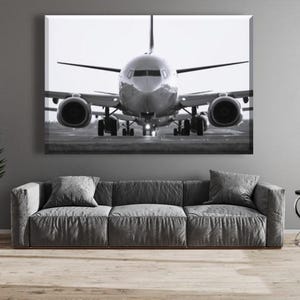 Könnte beinhalten: Schwarz-Weiß-Leinwanddruck eines Flugzeugs auf einer Start- und Landebahn, von vorne betrachtet. Das Kunstwerk hängt über einem grauen Ecksofa mit passenden Kissen. Eine hohe, dunkelgrüne Pflanze und ein kleiner Beistelltisch vervollständigen den Raum.