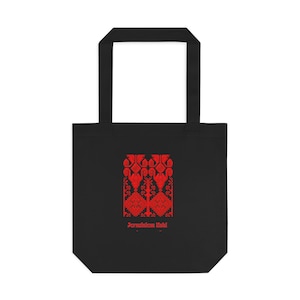 May include: A black tote bag featuring a red geometric pattern centered on the front. The design includes symmetrical shapes and the text Jerusalem Koki printed in red below the graphic. The bag has two black shoulder straps attached at the top.