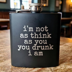 Könnte beinhalten: Schwarze Edelstahl-Taschenflasche mit silbernem Verschluss und Scharnier. Auf der Flasche steht der weiße Schriftzug "I'm not as think as you drunk I am". Sie liegt auf einem Holztisch.