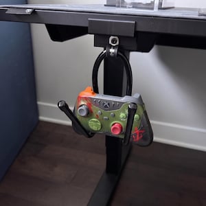 May include: A black metal desk-mounted controller hanger holding a gray and green gaming controller with red accents and a red logo. The hanger features a curved support arm and a clamp mechanism that attaches directly to the edge of a desk surface.