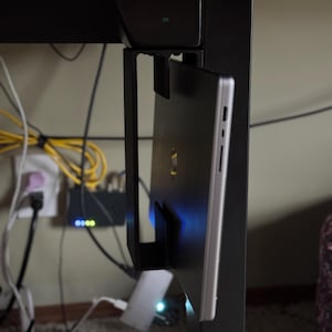 Pu&ograve; includere: Un supporto per laptop in metallo nero attaccato a una gamba di scrivania nera. Un laptop argentato &egrave; fissato nel supporto. Cavi elettrici e un router sono visibili sullo sfondo.