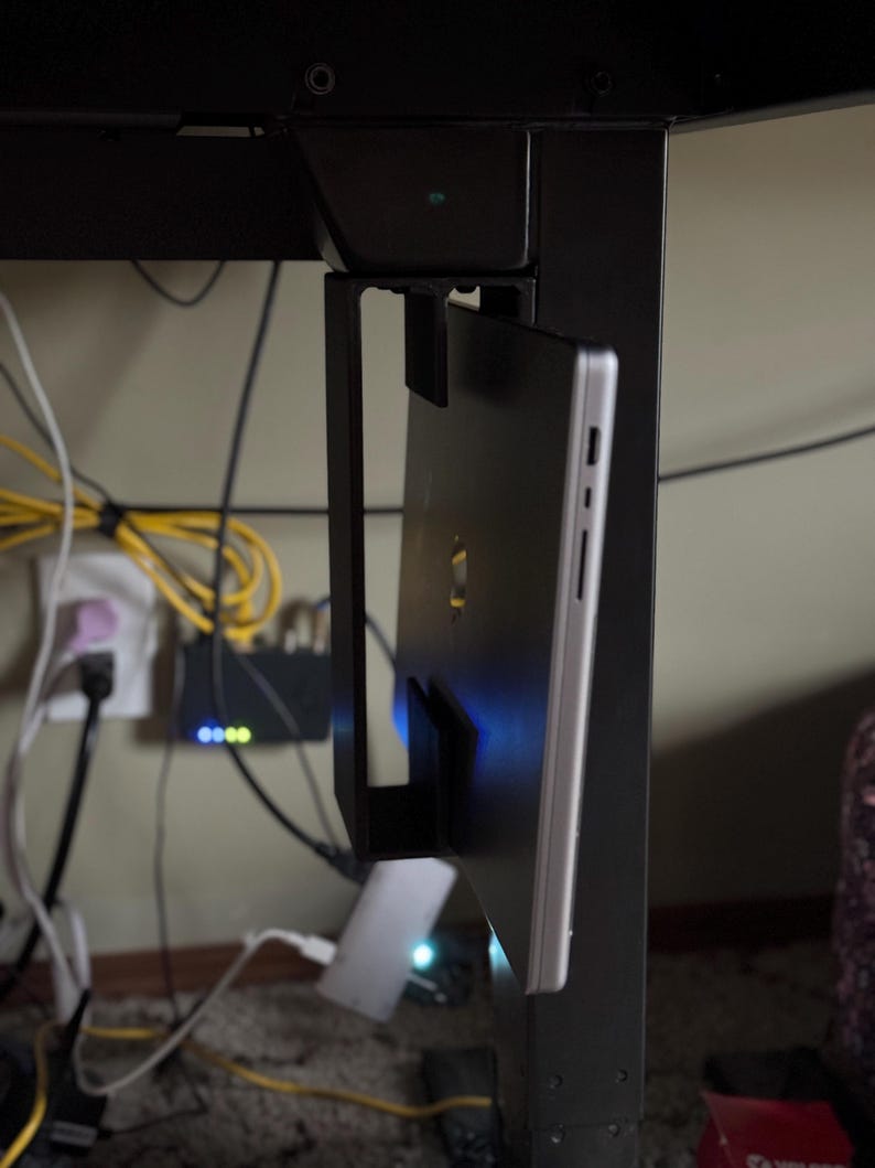 Pu&ograve; includere: Un supporto per laptop in metallo nero attaccato a una gamba di scrivania nera. Un laptop argentato &egrave; fissato nel supporto. Cavi elettrici e un router sono visibili sullo sfondo.
