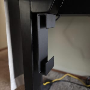May include: Close-up of a black metal desk accessory. The accessory is a bracket with a U-shaped design, mounted on the side of a black desk leg. The bracket has a hook-like extension. The background is a neutral wall and carpet.