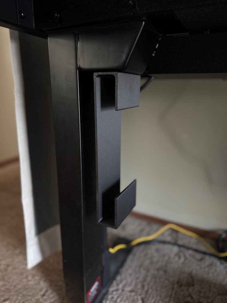 May include: Close-up of a black metal desk accessory. The accessory is a bracket with a U-shaped design, mounted on the side of a black desk leg. The bracket has a hook-like extension. The background is a neutral wall and carpet.