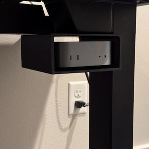 May include: A black, rectangular under-desk computer mount holding a silver computer. The mount is attached to a black desk frame. A white electrical outlet is visible on the wall below.
