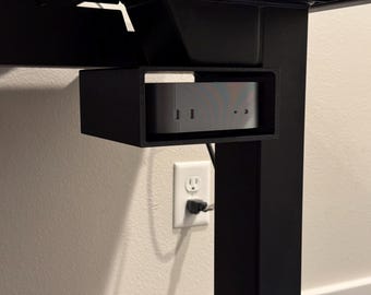 Puede incluir: Un soporte negro y rectangular para ordenador debajo del escritorio que sostiene un ordenador plateado. El soporte está fijado a un marco de escritorio negro. Un enchufe eléctrico blanco es visible en la pared de abajo.
