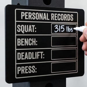 May include: Black rectangular sign with white text that reads "PERSONAL RECORDS". The sign has sections for squat, bench, deadlift, and press. The squat section shows "315 lbs" written in white. A hand is holding a white marker.