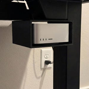 May include: A silver computer is mounted inside a black bracket under a black desk. The computer has three small indicator lights. An electrical outlet is visible on the wall.