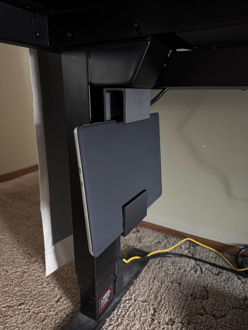May include: Black tablet holder attached to a black adjustable desk frame. The holder is designed to securely hold a tablet, keeping it within easy reach. The setup is ideal for a home office or workspace.