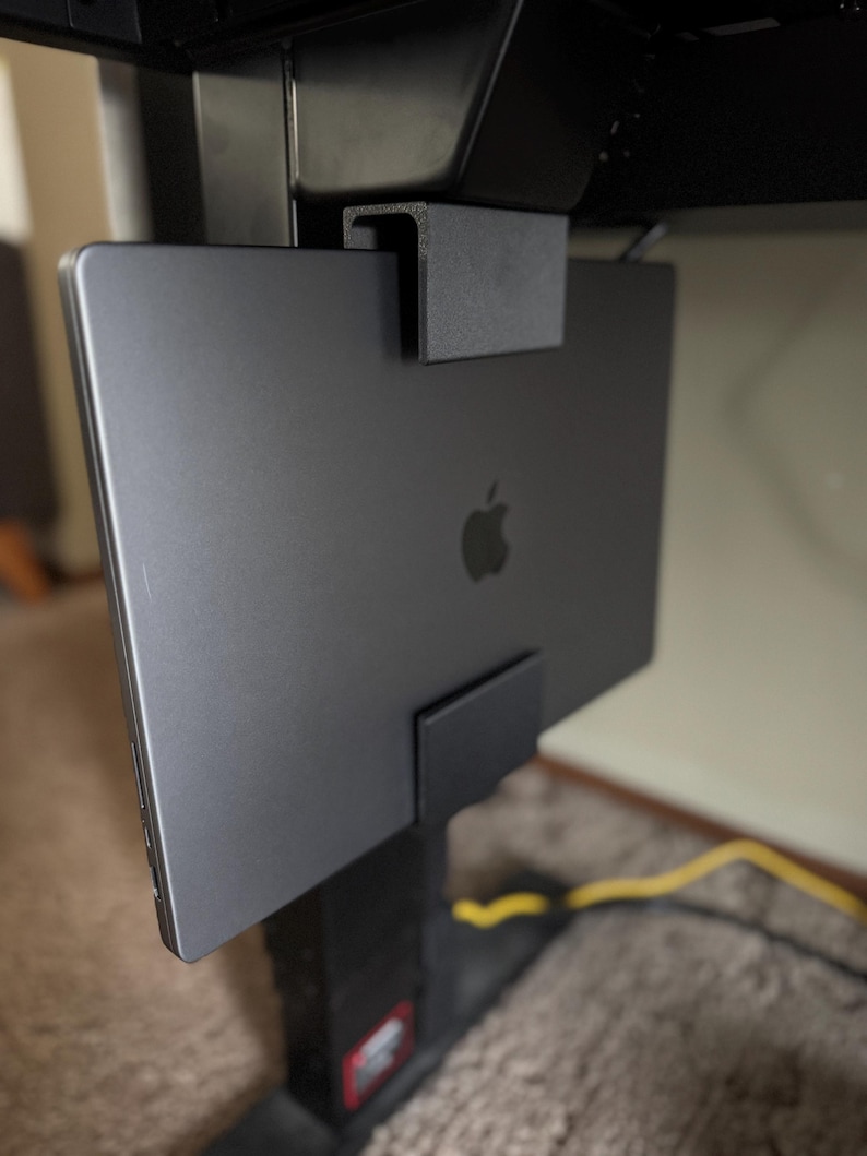 May include: A black laptop is mounted on a black metal bracket, attached to a black desk frame. The laptop is closed, displaying the Apple logo. The bracket appears to be designed for under-desk storage, keeping the laptop off the desk surface.