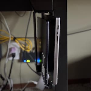 May include: A black metal under-desk laptop mount holding two laptops. One laptop is silver, the other is black. The mount is attached to a black desk leg. Cables and a network device are visible in the background.