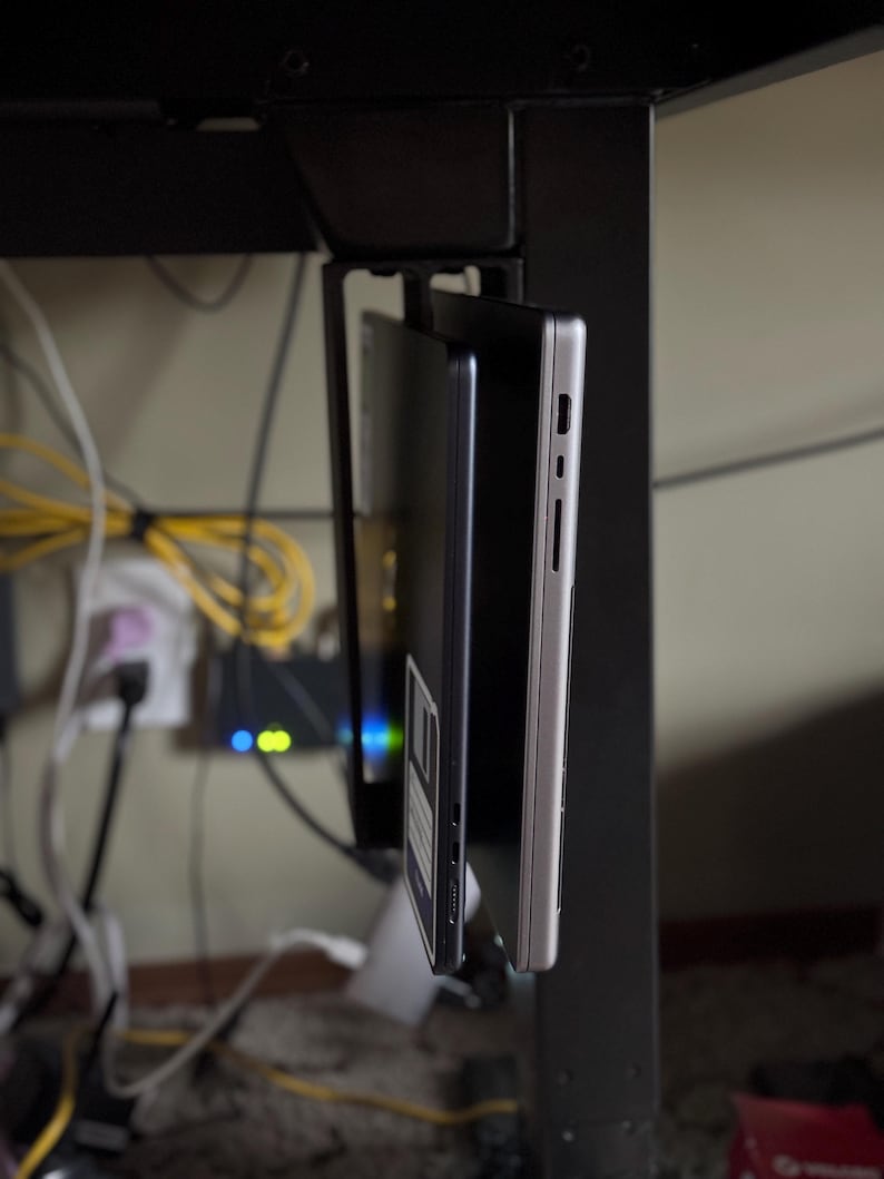 Pu&ograve; includere: Un supporto in metallo nero per laptop sotto la scrivania che contiene due laptop. Un laptop &egrave; argentato, l'altro &egrave; nero. Il supporto &egrave; attaccato a una gamba della scrivania nera. Cavi e un dispositivo di rete sono visibili sullo sfondo.