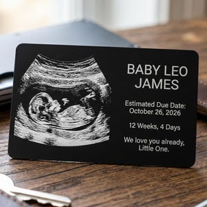 Peut inclure: Une carte rectangulaire noire avec une image d'échographie d'un fœtus. La carte porte l'inscription "BABY LEO JAMES" et la date d'accouchement estimée du 26 octobre 2026. Il est également indiqué "12 semaines, 4 jours".