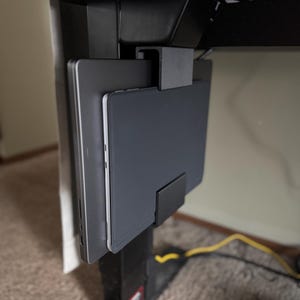 May include: A black under-desk laptop and tablet holder. The holder is mounted to the underside of a black desk and features a slot to hold a laptop and a tablet. The laptop and tablet are gray.