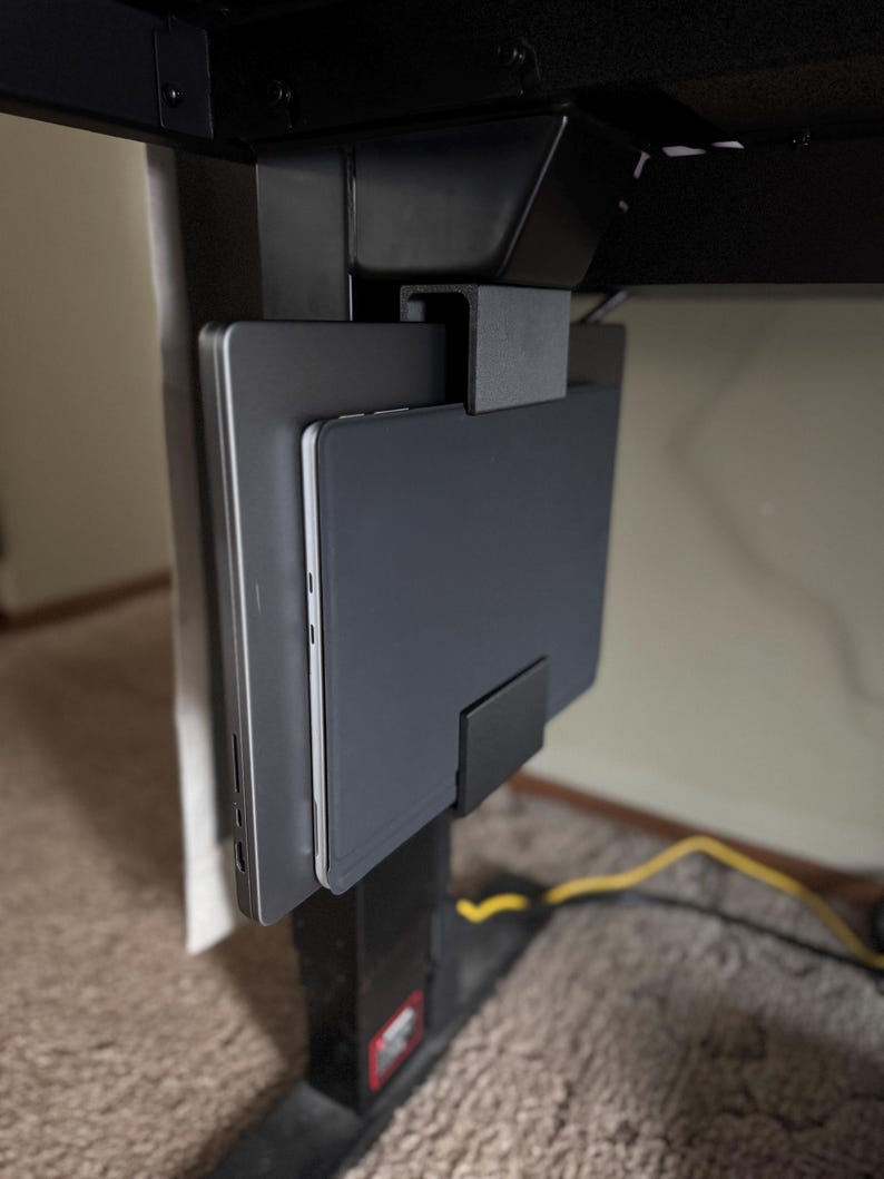 May include: A black under-desk laptop and tablet holder. The holder is mounted to the underside of a black desk and features a slot to hold a laptop and a tablet. The laptop and tablet are gray.