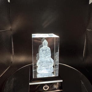 May include: A clear rectangular glass block featuring a laser-etched three-dimensional figure of a seated Buddha. The glass block rests on a black square base that includes a power button symbol on the front, illuminated by an internal light.