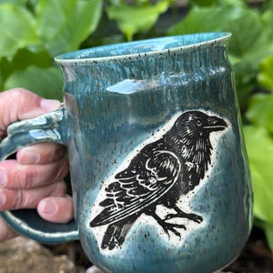 May include: A ceramic mug with a glossy teal and blue glaze featuring a speckled finish. The mug displays a black ink-style illustration of a raven on the side. It has a sturdy handle and a wide rim, showcasing a handcrafted appearance.