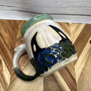 Może przedstawiać: Ceramiczny kubek z błyszczącym jasnozielonym brzegiem, który przechodzi w białe ściekające szkliwo na czarnej i głęboko niebieskiej teksturowanej podstawie. Kubek posiada zakrzywione ucho z ciemnoniebieskim wykończeniem oraz gładki, zaokrąglony kształt.