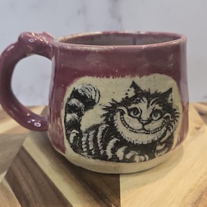 May include: A ceramic mug with a purple exterior and a smiling Cheshire Cat design. The mug has a rounded shape with a handle and is set on a wooden surface.