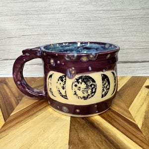 Op de afbeelding: Een handgemaakte keramische mok met een donkerpaarse buitenkant en een blauwe binnenkant. De mok heeft een ontwerp van de maanfasen in zwart en wit. Het handvat is ook donkerpaars.