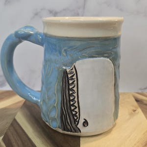 May include: Ceramic mug with a whale design. The mug features a light blue exterior with a sculpted wave pattern and a white whale illustration. The handle is also light blue. The rim is off-white.