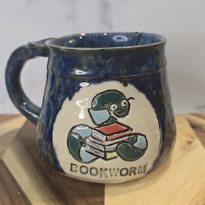 May include: A handmade ceramic mug with a blue exterior and a white oval design. The design features a cartoon bookworm holding books, with the word "BOOKWORM" below. The mug has a handle.