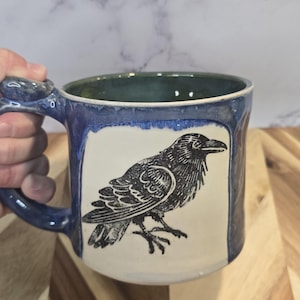 May include: A white ceramic mug with a black handle and the text "But First, Coffee" in a handwritten style. The mug is on a white surface. The mug is approximately 10 cm tall.